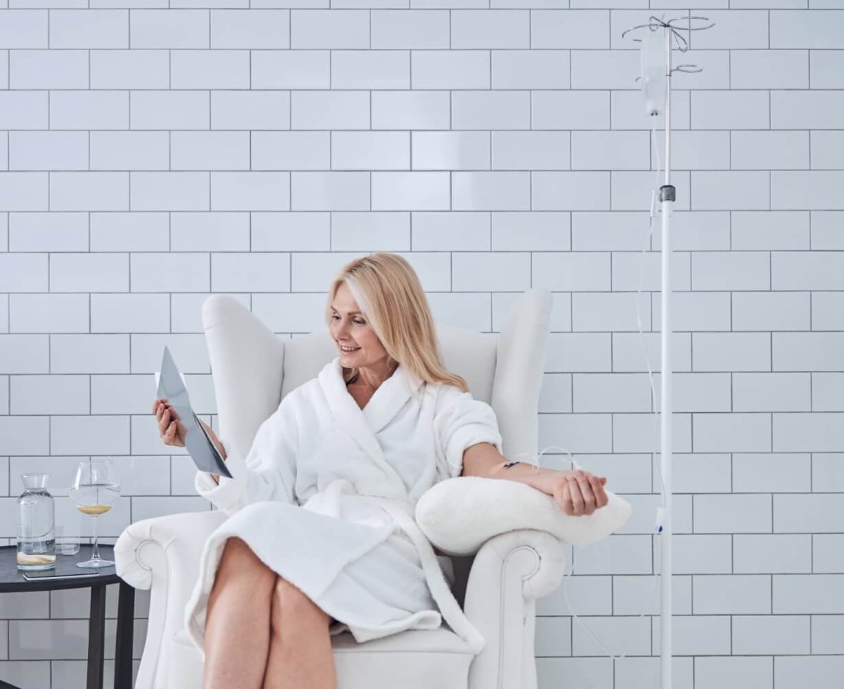 woman in bathrobe sitting on chair while receiving intravenous vitamin therapy in beauty center